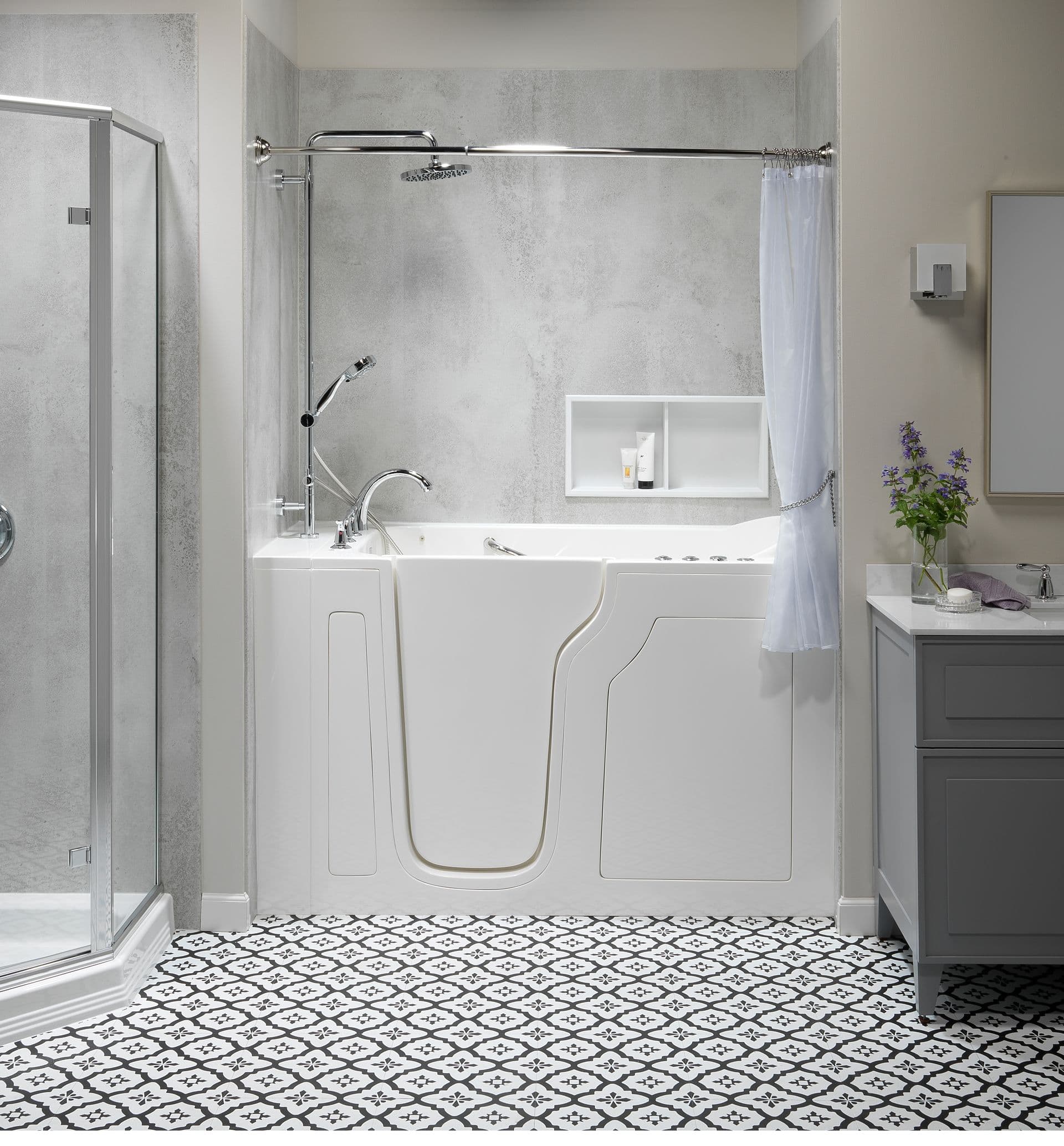 Bright, modern bathroom with white walk-in bathtub, gray tiles, geometric floor, glass shower, and vanity with purple flowers. Clean, calming atmosphere.