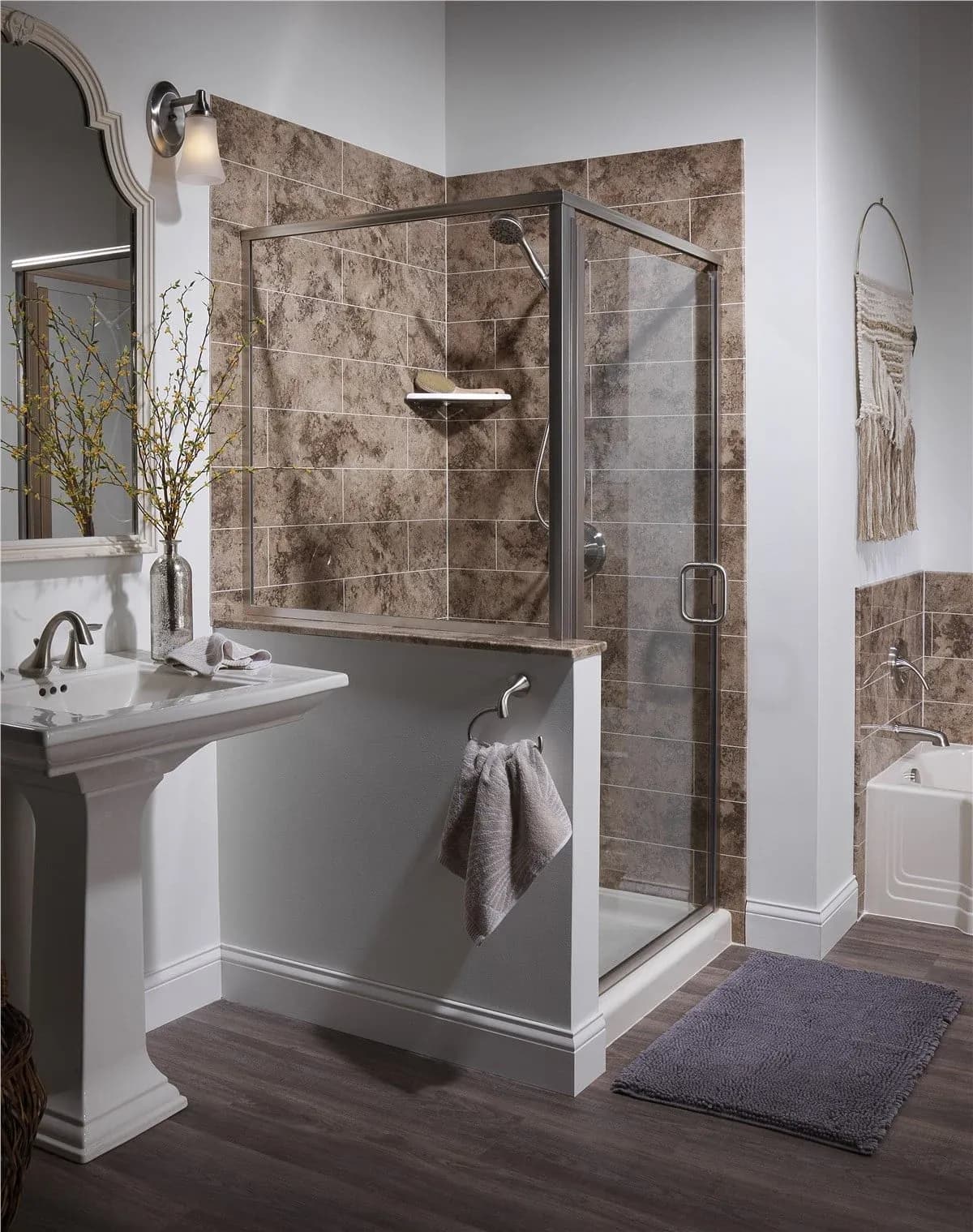 A modern bathroom features a glass-enclosed shower with brown tile, a white pedestal sink with a mirror and light, and a gray rug on a wooden floor.