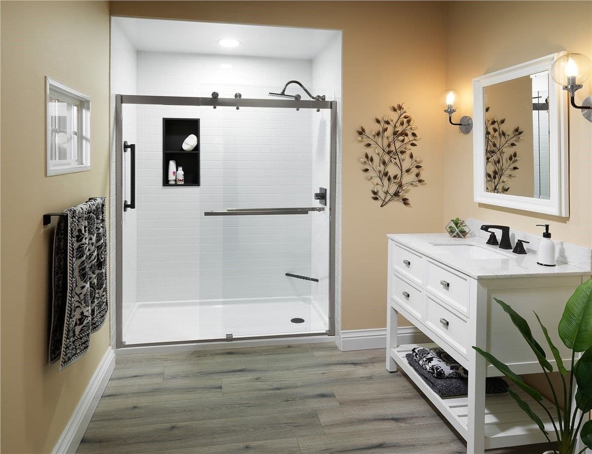 Modern bathroom with beige walls and wood floor features a white shower with glass doors. A white vanity with mirror, decorative plants, and black towel accents complete the elegant look.