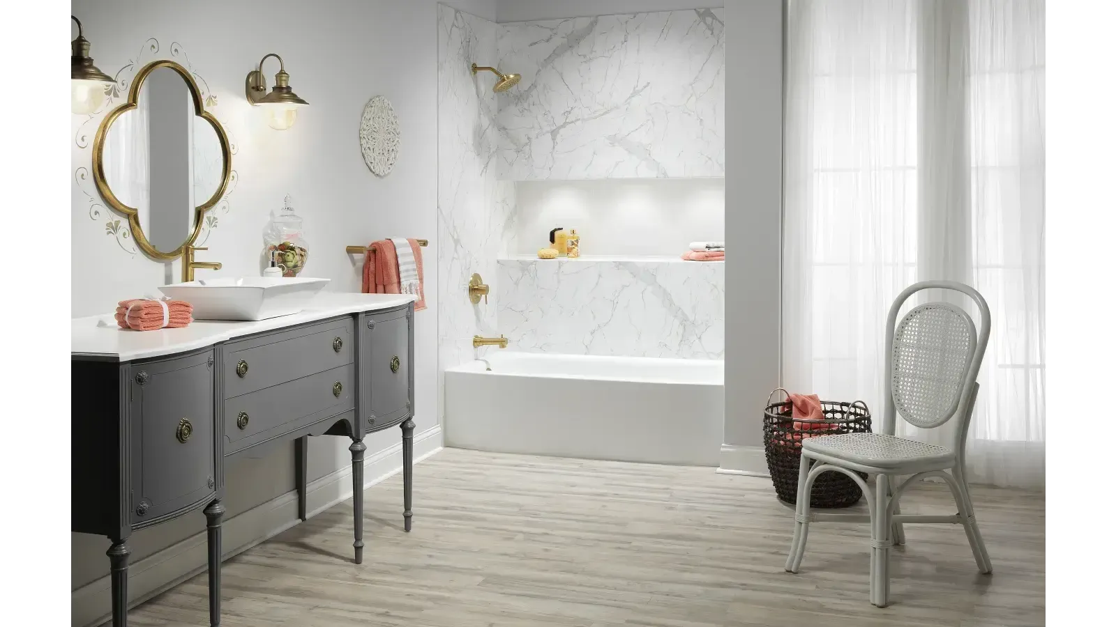 Elegant bathroom with a marble-tiled bathtub and gold fixtures. A vintage vanity with a mirror, wicker chair, and natural light create a serene ambiance.