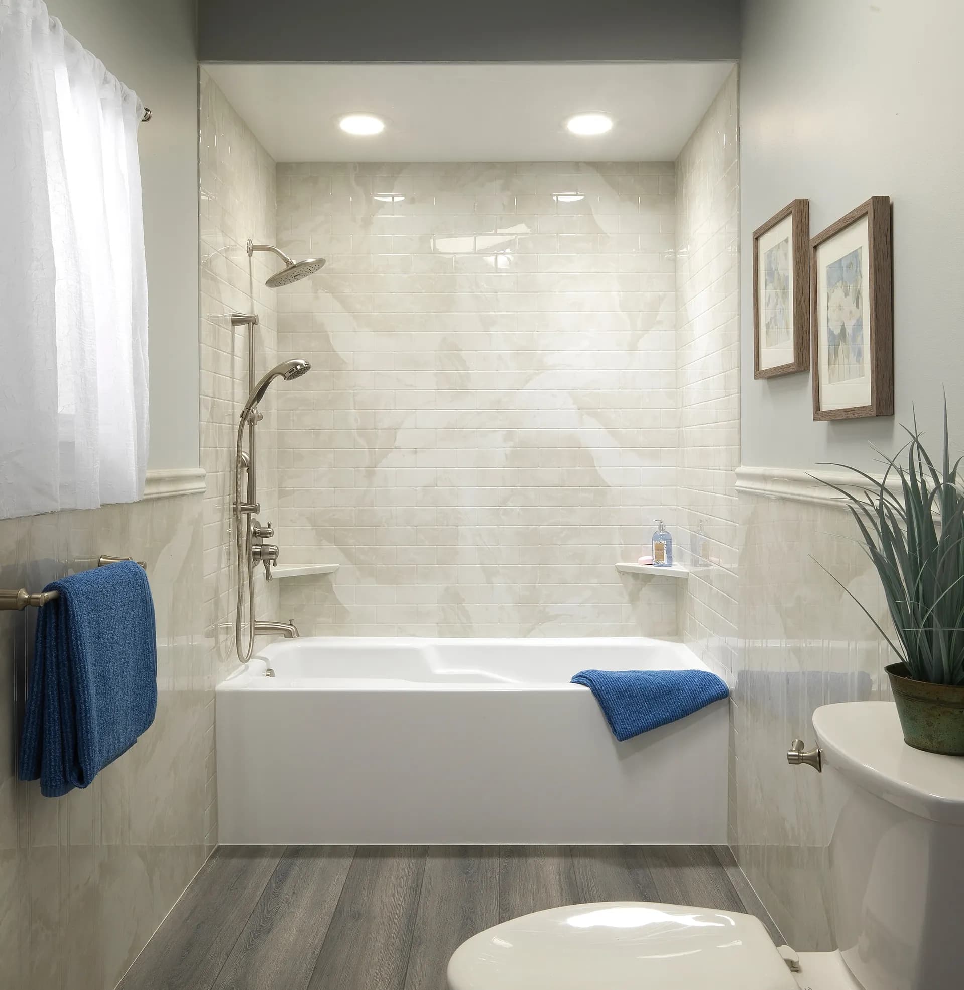 A modern bathroom with light beige marble walls, featuring a white bathtub with a shower and blue towels. Elegant décor includes framed art and a potted plant, creating a serene ambiance.