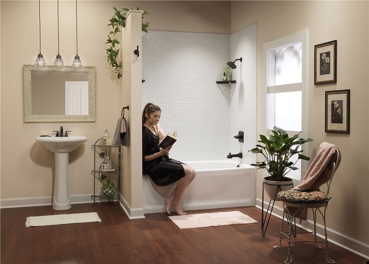 Woman in a black robe sits on the edge of a white bathtub, reading a book. The warm-toned bathroom features elegant lighting, plants, and decor.