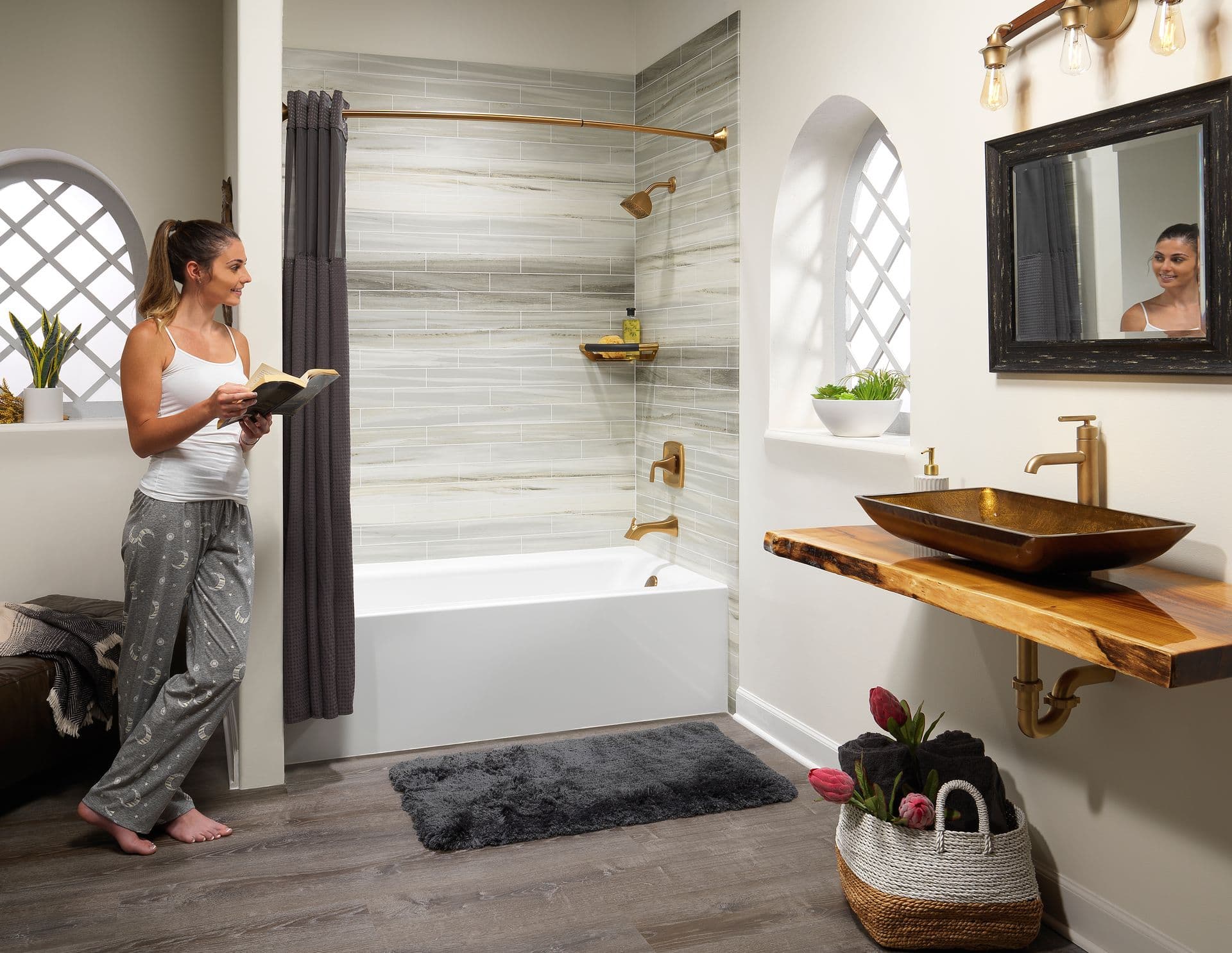 A woman in casual loungewear reads a book in a modern bathroom. The space features a tub with gray tiles, gold fixtures, a vessel sink, and soft lighting.
