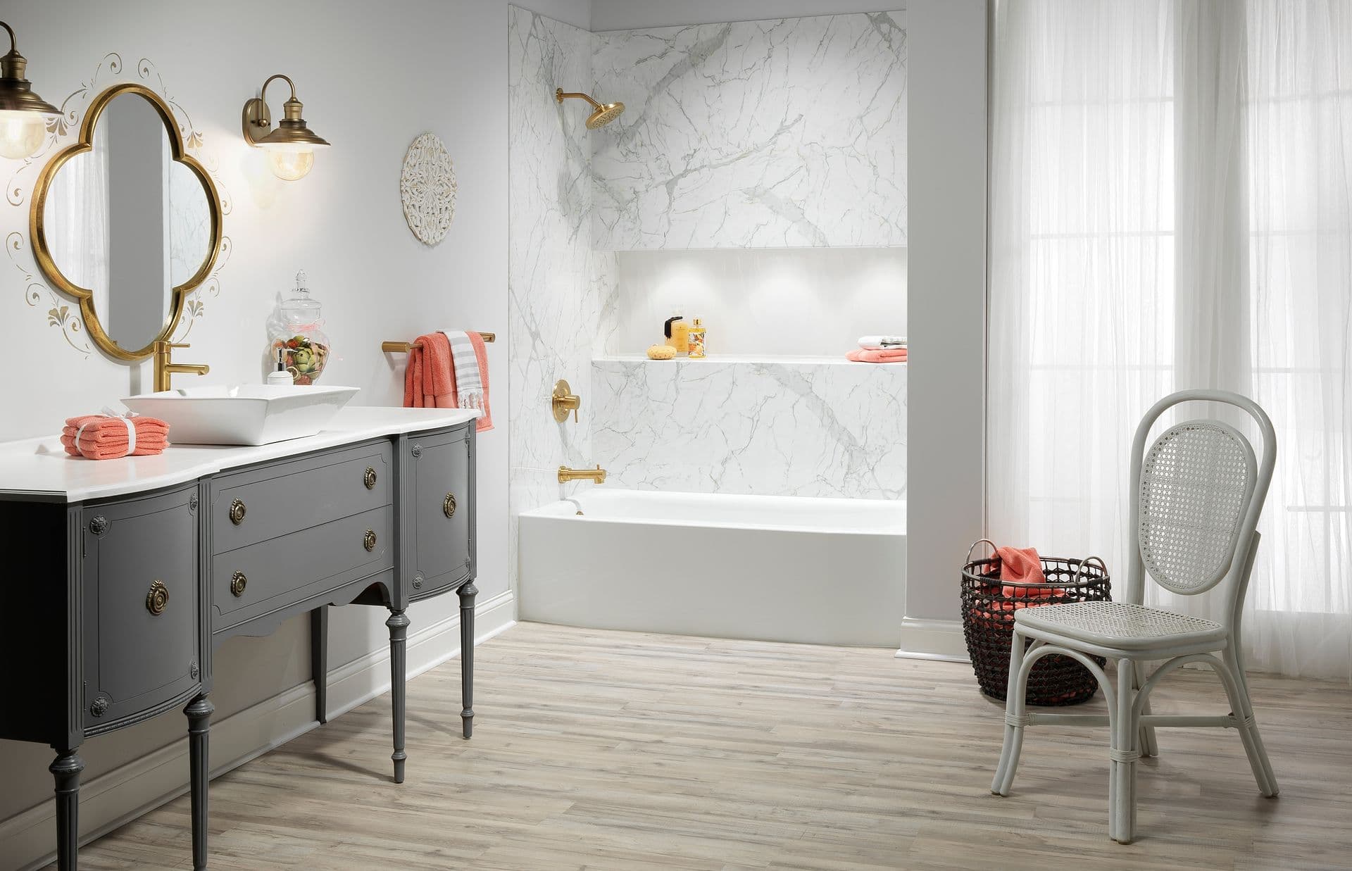 Elegant bathroom with a vintage gray vanity, white marble tub, gold fixtures, and soft lighting. A wicker chair and sheer curtains add cozy charm.