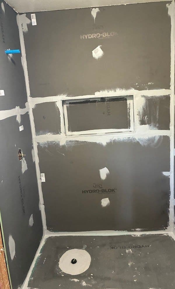 Unfinished shower area with grey waterproof walls, seams sealed with white tape. A niche and step are visible. Mood is under-construction.